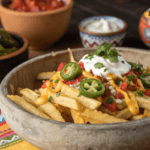 A bowl of Papas Locas topped with melted cheese, guacamole, sour cream, tomatoes, and jalapeños on a colorful tablecloth